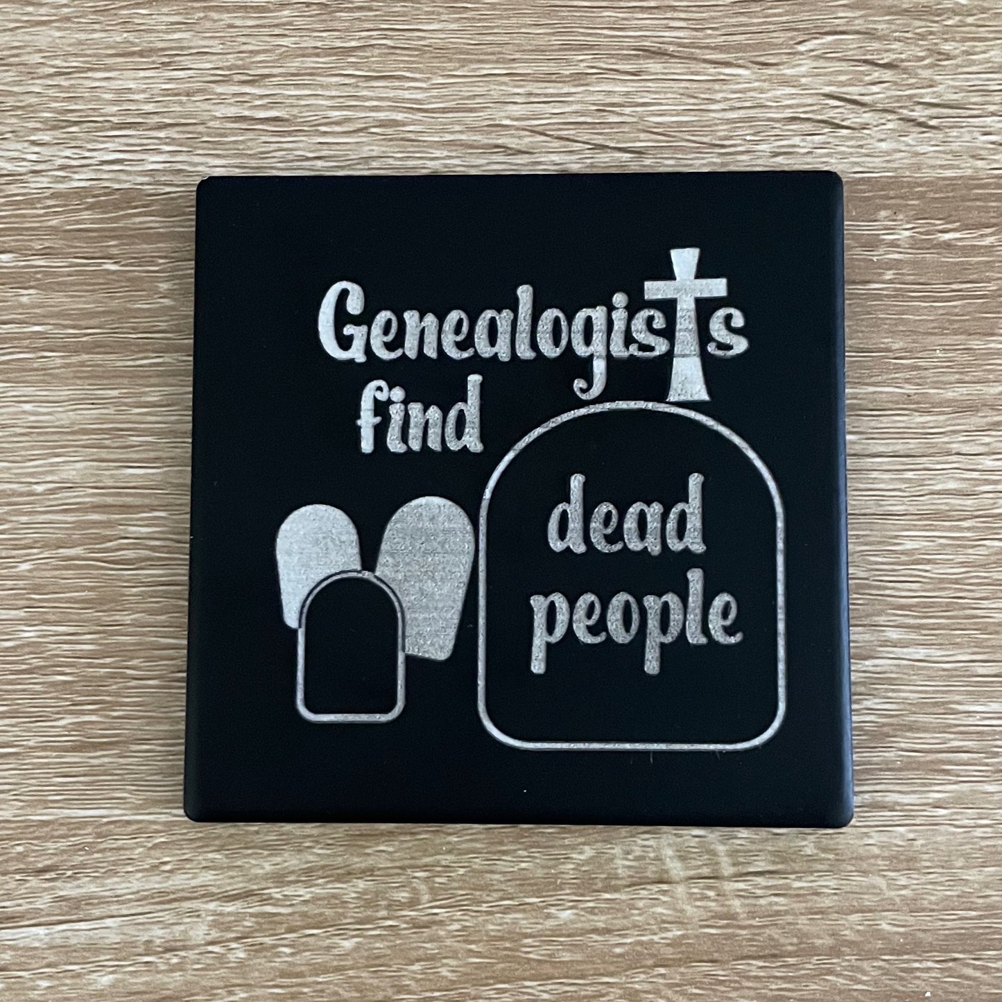 Black ceramic square coaster engraved with Genealogy phrase "genealogists find dead people" and gravestones