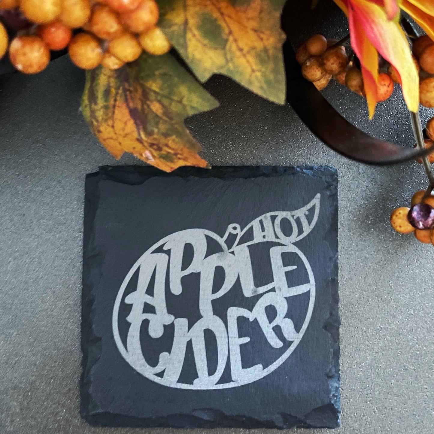 Square black slate coaster laser engraved with hot apple cider text inside of apple shape. Coaster is surrounded by fall decor.