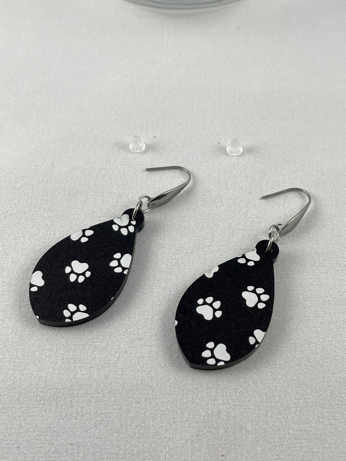 Detail view of Black acrylic teardrop shaped dangle earrings with white paw prints and stainless steel ear wires with teardrop accent and included silicone earring backs