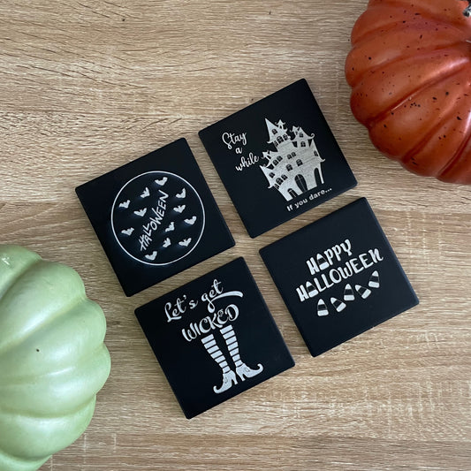 Set of four black square ceramic coasters engraved with Halloween designs including bats, haunted house, candy corn and witch's striped socks and heels